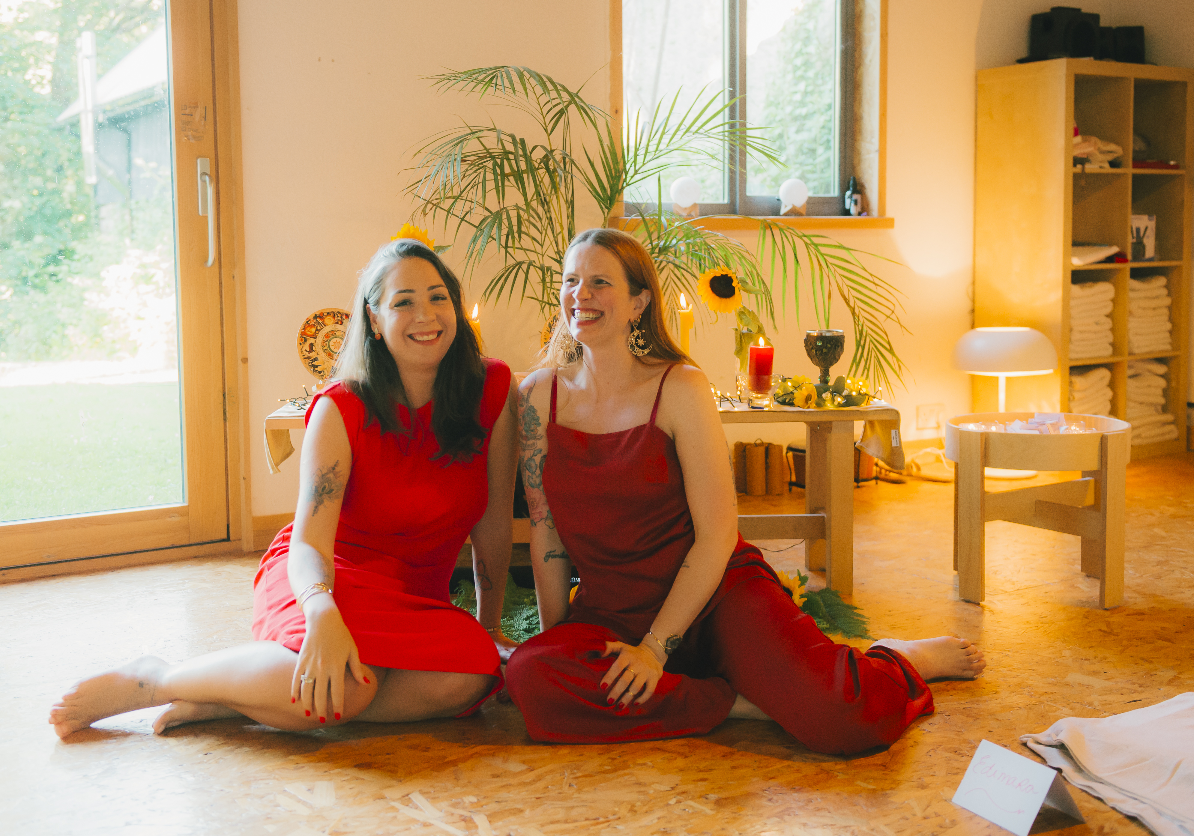 Bruna e Katita sorrindo, sentadas juntas em frente a um altar com velas e girassóis